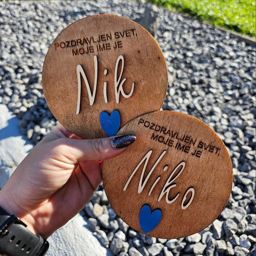 Two wooden coasters with engraved names and a blue heart held by a hand on a gravel surface.